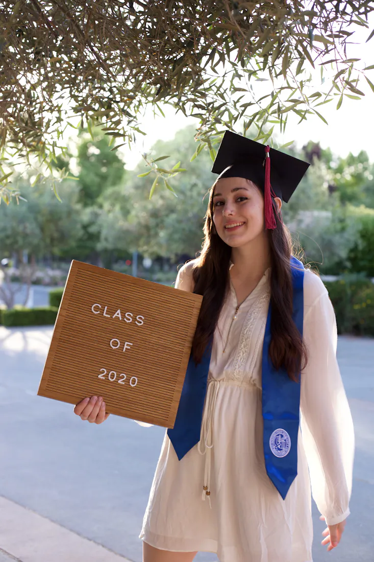A graduate in cap and gown holding a class sign